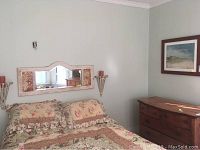 Bedroom wall featuring rectangular mirror above bed with floral bedspread, two metal wall sconces with red candles on each side of the mirror, and framed art above dresser.