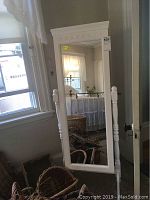 White framed floor mirror standing on two supports showing carved decoration and dimensions near window.
