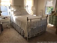 Front angled view of the full metal bed frame with white finish and brass top rail and finials, with bedding and pillows present but not included.