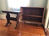 Photo showing the wooden bookcase and ornate wood table placed side by side on a wood floor near a wall with a window.