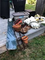 Full view of lot showing plant stand, terracotta pots, sea shell planter, gloves, hand tools, sprinkler, and hose with box.