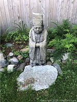 Full view of cement Asian statue standing outdoors in garden bed with plants and rocks nearby.