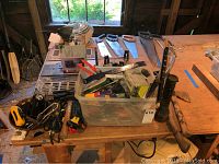 Wide view of various hand tools laid out on a wooden workbench and inside a plastic bin in a barn environment.