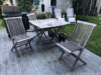 Full view of teakwood table and three matching chairs on deck, showing weathered condition and outdoor environment.