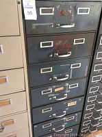 Front view of gray metal storage cabinet with seven drawers, showing label holders and handles with some faded writing on the drawers.