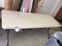 View of the entire folding table showing beige plastic tabletop and black legs in garage setting.