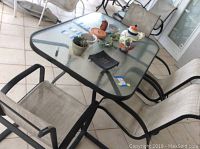 Patio dining table with glass top and four beige mesh sling chairs arranged around it.
