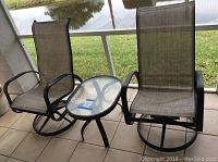 Two black metal frame rocking patio chairs and one oval glass-top side table placed on a tiled patio with grass and water in the background.