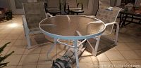 Patio set showing round glass-top table with two white metal chairs on tiled floor.