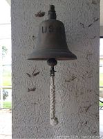 Full front view of the solid brass bell with USN embossed and braided rope pull hanging from the clapper, mounted on textured wall.