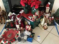 Large photo showing assortment of Christmas decorations on tiled floor including various Santa figurines, poinsettias, LED lights, storage crate, quilt, and plates.
