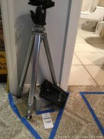 Silver tripod standing with legs extended. Blue painter's tape on carpet around it and a MaxSold tag labeled 144 at base. A black pouch with Giotto logo is placed at the tripod base.