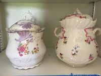 Two vintage porcelain chocolate pots side by side on a shelf. The left pot has lavender and gold accents with floral design; the right pot has pink-red accents with floral design and two handles.