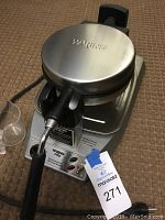 Front perspective photo of Waring Pro waffle maker showing overall shape, stainless steel body, and attached power cord with a batter measuring cup next to it.