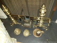 Photo showing two brass andirons, two metal kettles, square decorative metal vase, metal mug, and candle snuffer arranged on floor in front of fireplace screen.