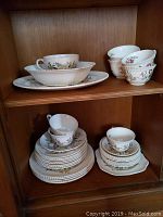 Display of stacked plates, cups and serving pieces in a wooden cabinet with floral design on china.