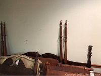 Photo showing a variety of bed frame parts against a wall including the king bed frame's arched header and footboard and the tall posts of the queen bed frame.