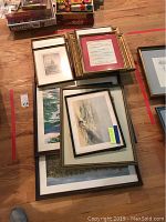 Grouped collection of framed art prints and sketches stacked on the floor, some with red, gold, and black frames, sizes vary.