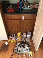 Wide view showing a wooden wall cabinet under a sink with various tools on the countertop and a collection of cords, light bulbs, and accessories on the floor.