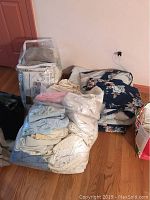 Photo showing bags of various linens including sheets and pillow cases in plastic bags along with folded quilt and curtains on floor.