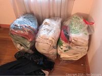 Three large clear plastic bags filled with assorted towels and blankets, placed against a wall near window curtains.