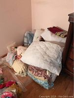 Photo showing assorted folded quilts and blankets stacked in a corner by wooden furniture on hardwood floor.