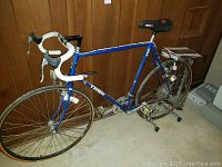 Full view of the blue Trek road bike mounted on a stationary stand, showing the frame, handlebars with white tape, wheels, and black Vettagel seat.