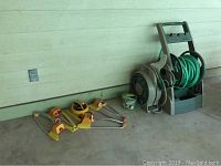 Photo showing two garden hoses on plastic reels and four yellow-orange plastic lawn sprinklers placed on the ground next to a wall.
