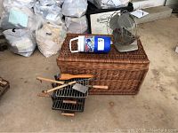 Photo of wicker basket, thermos, vintage fan, and hibachis with grill tools arranged on concrete floor.