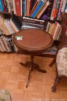 Full view of the round wood table with leather top, positioned by a bookshelf and a chair.