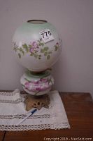 Full view of the lamp showing the round painted glass shade with pink flowers and green leaves, sitting on a lace doily.