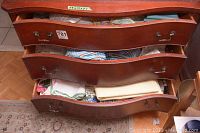 Three open wooden drawers containing assorted folded linens, including tablecloths, tea towels, dollies, and fabric pieces.