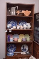 Wood cabinet shelf with 6 pastel coffee cups and saucers on bottom shelf, creamers and tea pot in middle, and decorative blue and white plates on top shelf along with wooden donkey figure.