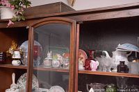 Wood framed glass cabinet filled with assorted glassware, china cups, plates, creamers, and decorative items.