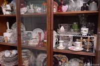 Wide view showing a wooden cabinet with multiple shelves containing assorted china plates, cups, glassware, and decorative items including figurines and painted eggs.