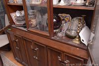 Wood cabinet with glass doors holding plates, cups, bowls, and decorative glass pitcher inside and several decorative items on top including plates and brass candle holders