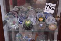 Overall view of shelf displaying twelve glass paperweights behind glass cabinet door