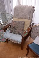Wooden armchair with beige throw and two cushions on seat and backrest. Shows wear on wood and cushions.