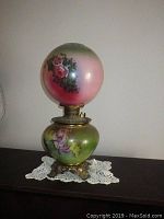 Front view of electrified oil lamp showing hand-painted rose motifs on globe shade and cherub designs on reservoir, with ornate metal base on a lace doily.