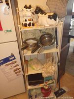 Photo of metal shelving unit holding ceramic cow cookie jars on top shelf, pots and pans on second shelf, and various kitchen items on lower shelves. Shows shelving size and condition.