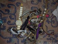 Photo shows a pile of assorted wristwatches with various metal and leather bands on a patterned carpet.