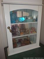 White wall mounted cabinet with glass door showing three shelves filled with various miniatures including blue perfume bottle, glass heart, decorative apple, trinket box, miniature squirrel, damaged glass animal, metal lantern ornament and pencil sharpener, miniature shoes, metal figurines including damaged and good Lesley items