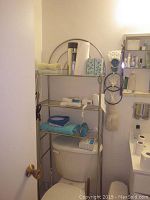 Metal over-the-toilet bathroom rack installed above white toilet, showing contents including towels, tissue box, paper towel roll, toiletries, and handheld shower head.