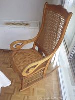 Wood cane rocking chair with visible cane seat and backrest, and wooden curved arms, set on parquet floor.