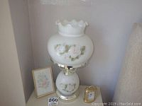 White glass lamp with floral painted roses, gold metal base, small Bulova clock, and framed floral art leaning against wall.