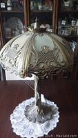 Full view of the metal table lamp showing the ornate shade with embossed floral and scroll details, metal finial on top, and electrical cord at base.