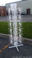 Full vertical view of the white steel multi-card holder stand placed outdoors on asphalt and grass.