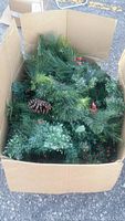 Photo of open cardboard box filled with artificial Christmas grouped greenery featuring pine needles, pine cones, and red berries.