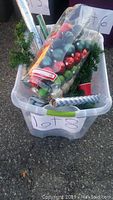 Plastic bin filled with Christmas decorations including multiple packs of colorful ornaments, green artificial garland, and rolls of gift wrap paper.