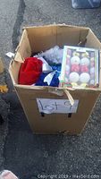 Large cardboard box labeled Lot 9, filled with assorted Christmas decor items including a boxed set of Christmas ornaments, a red Santa hat, silver tinsel, and other decorative pieces visible inside the box.